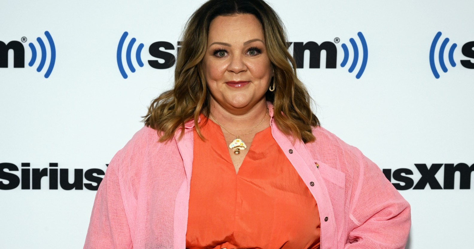 Cropped head and chest image of Melissa McCarthy who has spoken up against Tennessee Drag ban. She is wearing a orange top with a pink cardigan on a white background with Sirius XM logo.