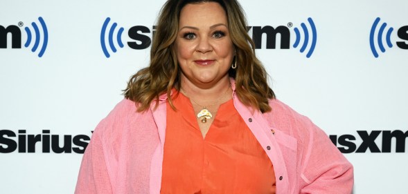 Cropped head and chest image of Melissa McCarthy who has spoken up against Tennessee Drag ban. She is wearing a orange top with a pink cardigan on a white background with Sirius XM logo.