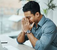A man is wearing a blue shirt with the sleeves rolled up to his elbow. He appears stressed as he has his hands in his face.