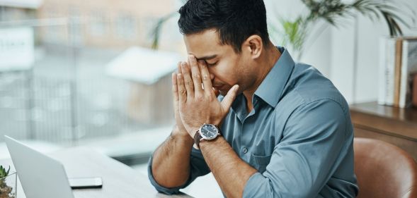 A man is wearing a blue shirt with the sleeves rolled up to his elbow. He appears stressed as he has his hands in his face.