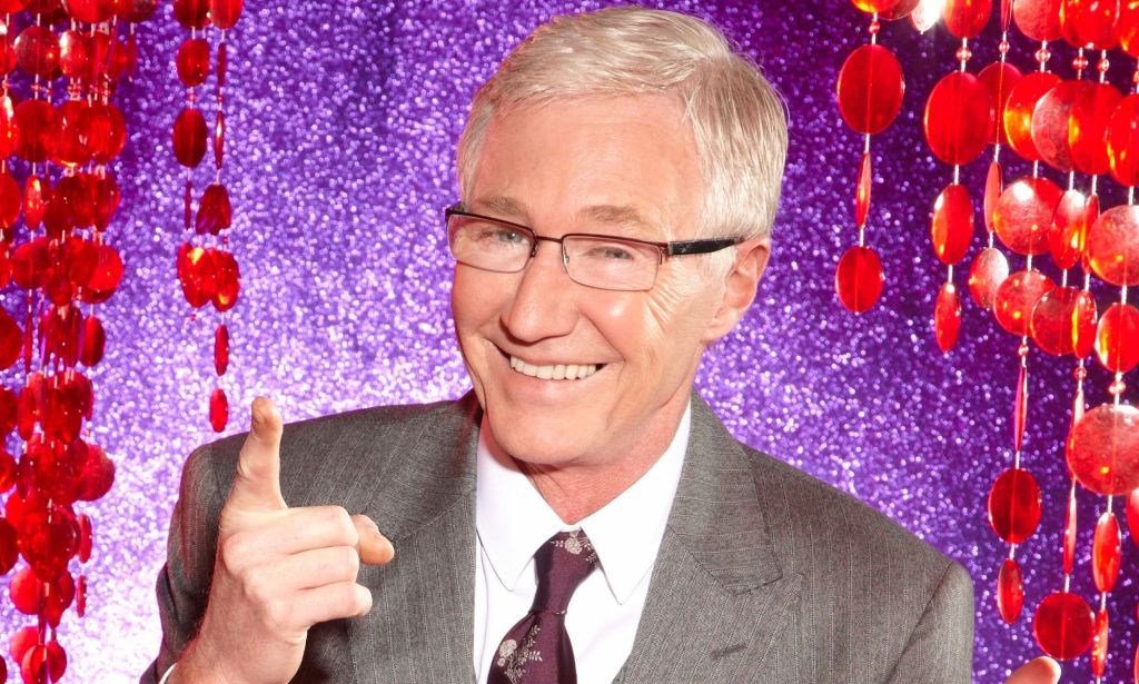 Paul O'Grady in a grey suit and tie, smiling and pointing upwards, against a purple and red background.