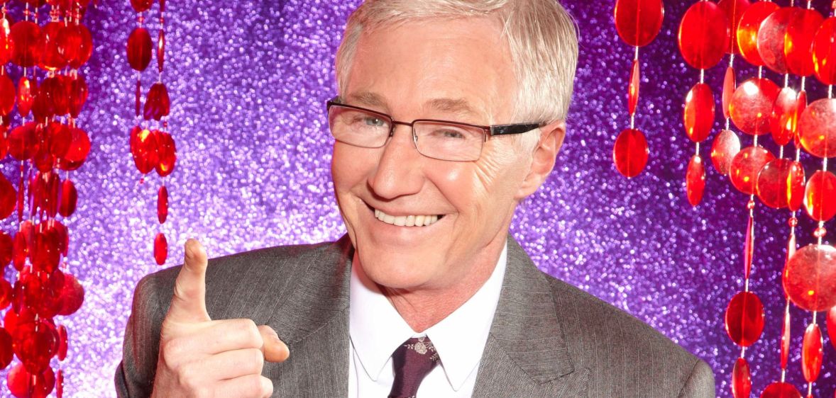 Paul O'Grady in a grey suit and tie, smiling and pointing upwards, against a purple and red background.