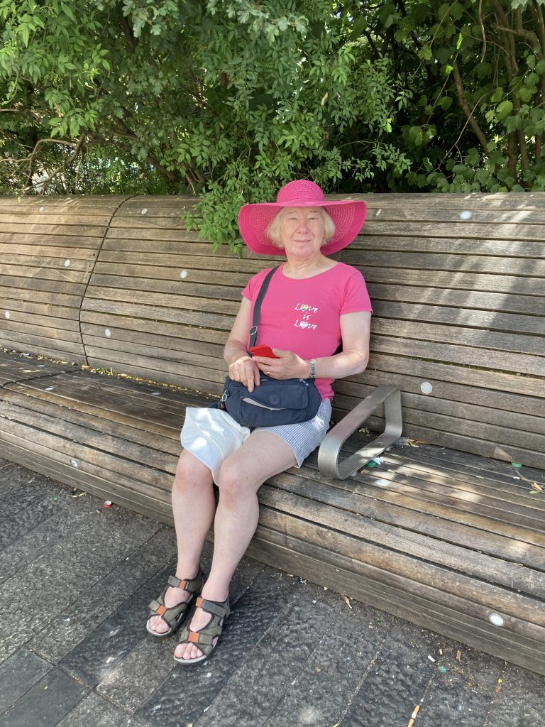 Petra is sitting on a wooden bench. She is wearing a pink had and pink tshirt and denim shorts. 