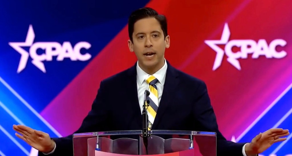 Daily Wire host Michael Knowles stands in front of a red and blue background reading "CPAC"
