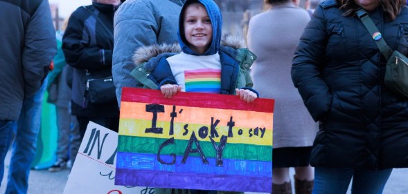 A child in a rainbow T-shirt holds a sign reading "It's okay to say gay"