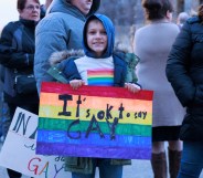 A child in a rainbow T-shirt holds a sign reading "It's okay to say gay"