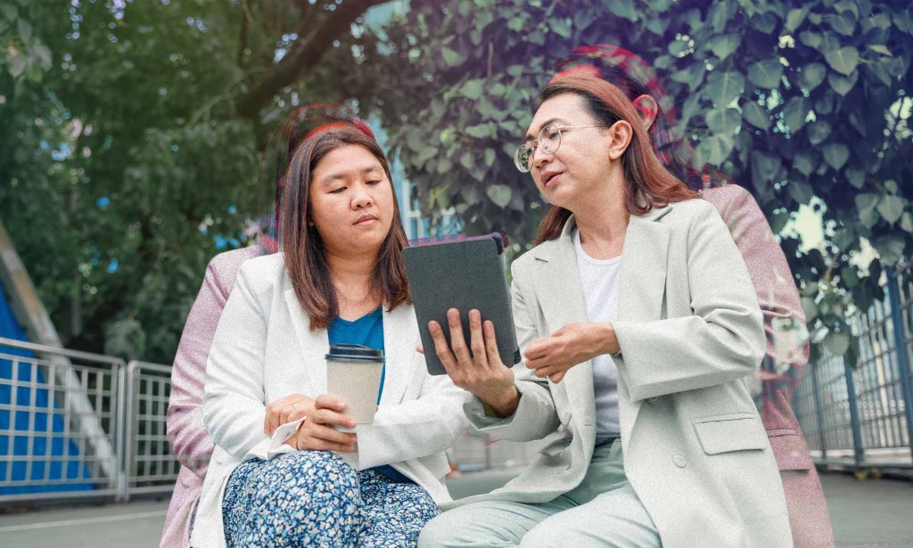 Two women are sitting outside. One woman is showing something on a tablet while the other woman looks on while holding a cup of coffee.