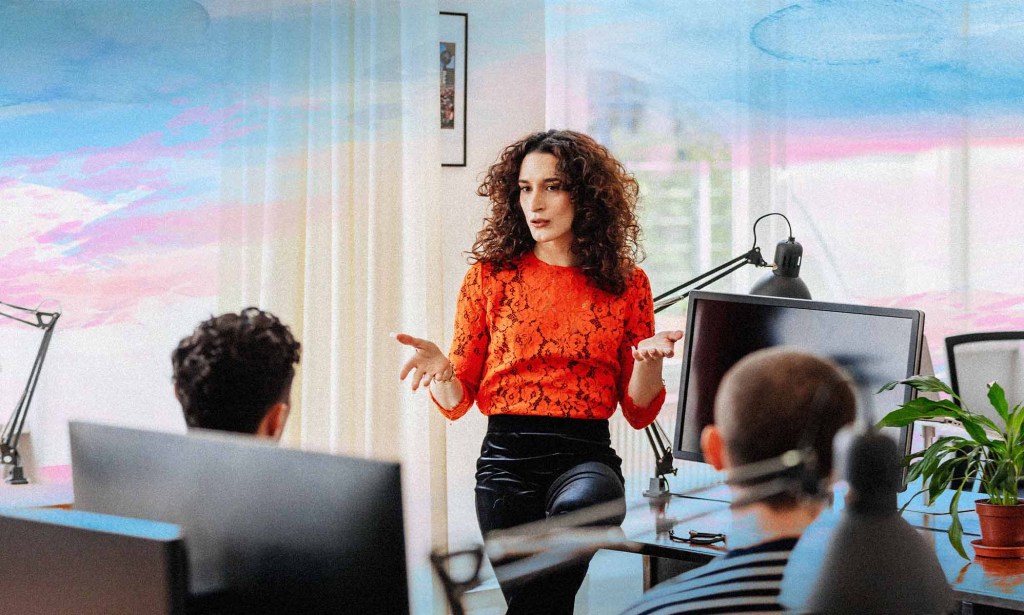 A person speaking in an office with trans Pride flag colours superimposed behind