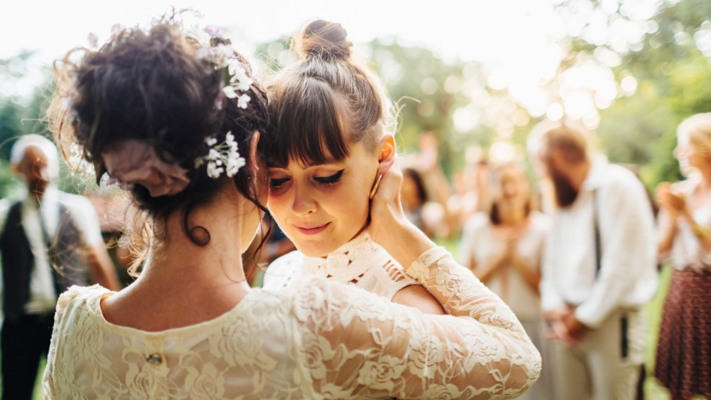 Two women embrace each other, both wearing wedding dresses