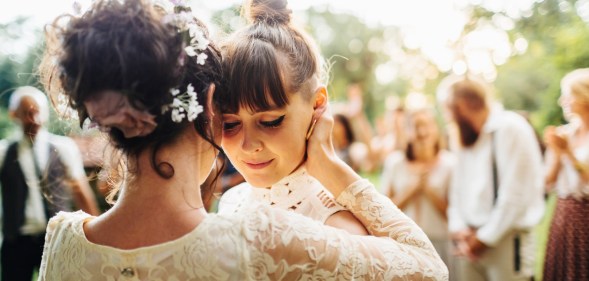 Two women embrace each other, both wearing wedding dresses