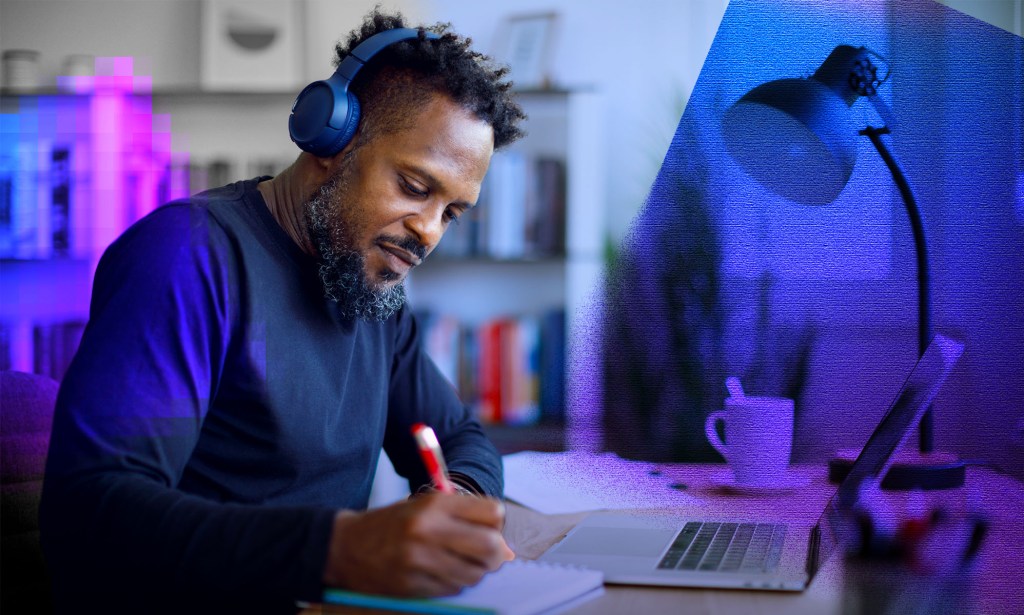 A mean is wearing a blue jumper and writing notes on a pad in front of his computer. 