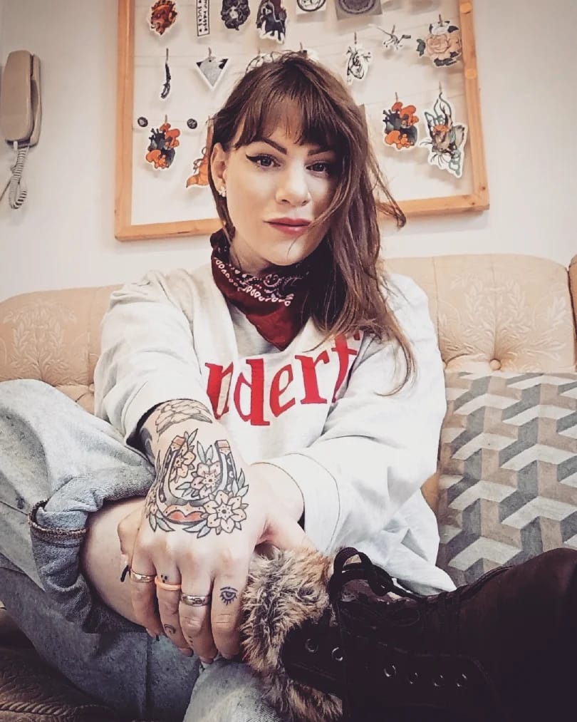 Victoria is posing for the camera, she is wearing a white jumper with a bandana and has colorful and vivid hand tattoos.