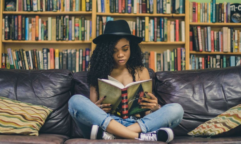 A woman wearing a black hat, blue jeans and black trainers reads a book on a black sofa against a backdrop of books.