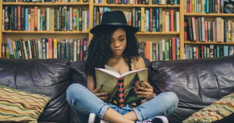 A woman wearing a black hat, blue jeans and black trainers reads a book on a black sofa against a backdrop of books.