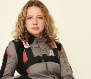 Queer stunt driver and professional drift driver Zandara Kennedy wears a grey, black and red motorsports outfit as she poses for the camera