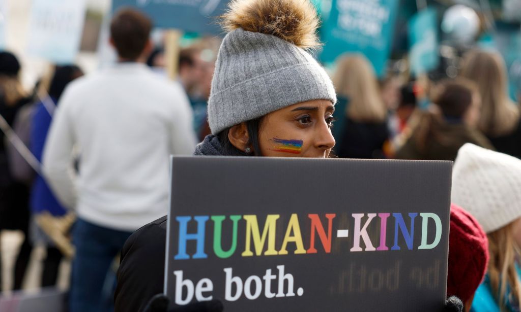 A person holds a sign reading 'Human-kind, be both' written in rainbow script at an LGBTQ+ rights protest