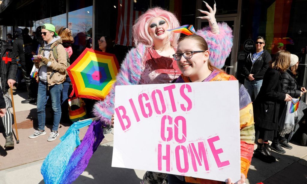 A person holds up a sign reading "Bigots go home" as a drag queen poses in the background at a protest in support of Drag Story Hour