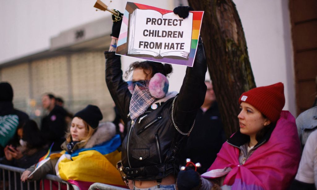 A person holds up a sign that reads "Protect children from hate" with a graphic of the Progressive Pride flag amid a crowd of LGBTQ+ people and allies counter-protesting against hate towards a drag event