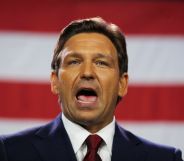Florida governor Ron DeSantis wears a suit and tie as he speaks with his mouth wide open. The red and white stripes of the US flag can be seen in the background
