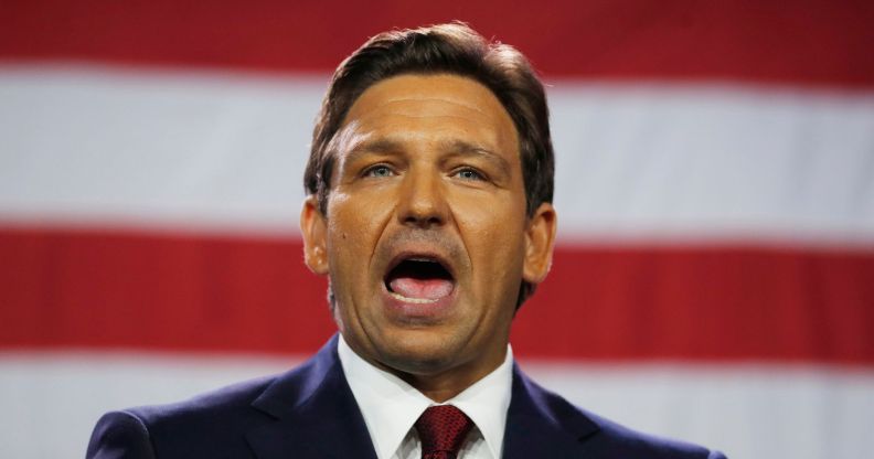 Florida governor Ron DeSantis wears a suit and tie as he speaks with his mouth wide open. The red and white stripes of the US flag can be seen in the background