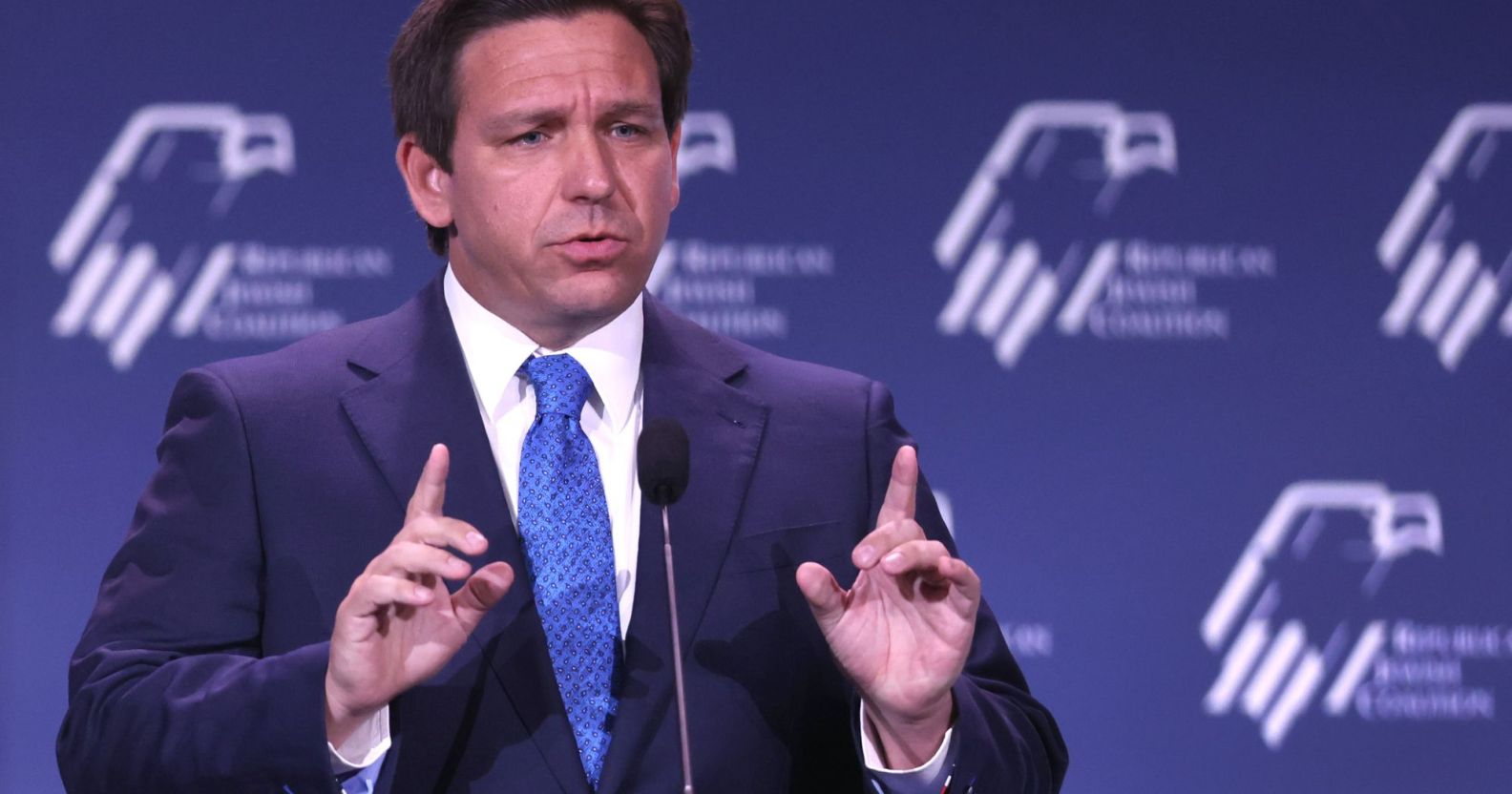 Florida governor Ron DeSantis wears a suit and tie as he gestures with one finger raised upwards on both hands during a speech