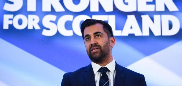Newly-appointed SNP leader Humza Yousaf wears a suit and tie as he stands in front of a sign in the colours of the Scottish flag
