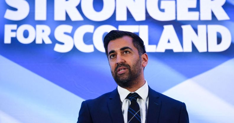 Newly-appointed SNP leader Humza Yousaf wears a suit and tie as he stands in front of a sign in the colours of the Scottish flag