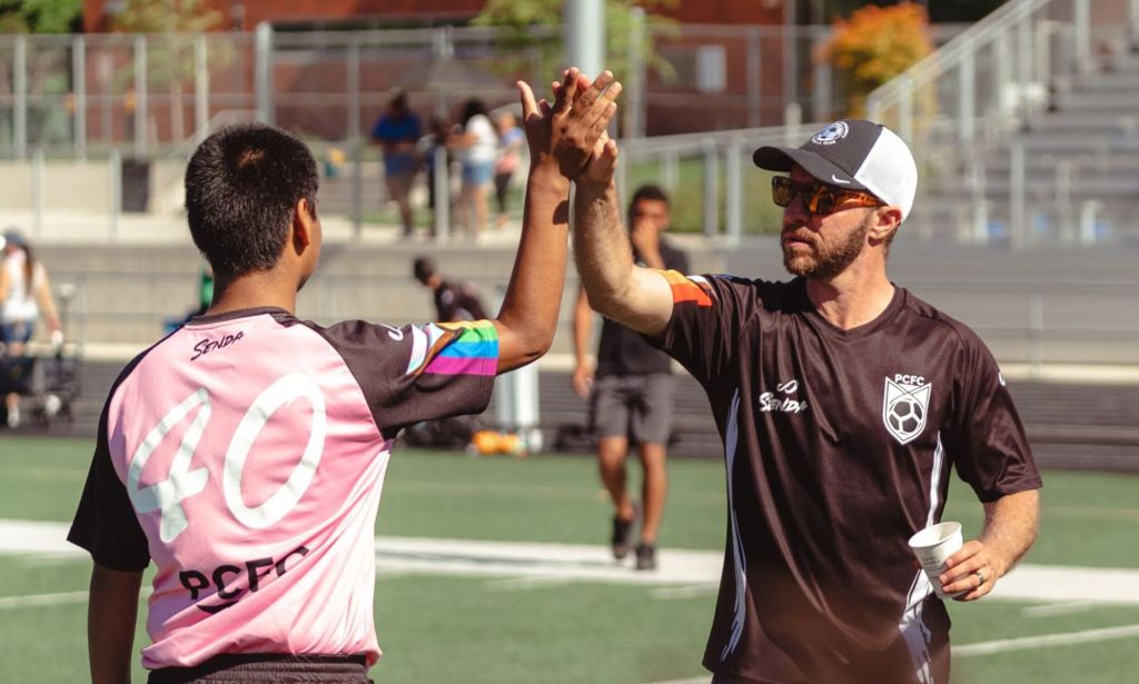 Openly trans coach Kaig Lightner wears a black football jersey as he high fives a person wearing a pink jersey on a sports field