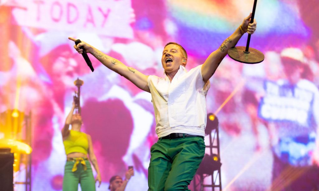 American rapper Macklemore wears a green and white outfit as he performs on stage with a rainbow LGBTQ+ flag seen in the background