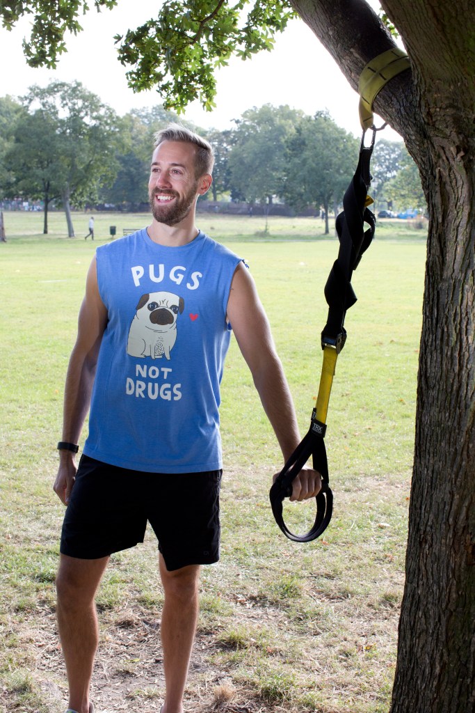 Matt Boyles pictured outdoors. He is wearing a shirt that says "pugs not drugs" and is standing by a tree with exercise equipment.