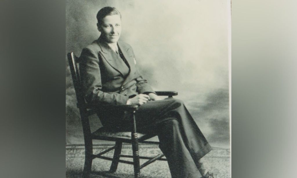 A picture of trans trailblazer Michael Dillon from 1944 where he's wearing dark slacks and a blazer while sitting in a chair with his legs crossed