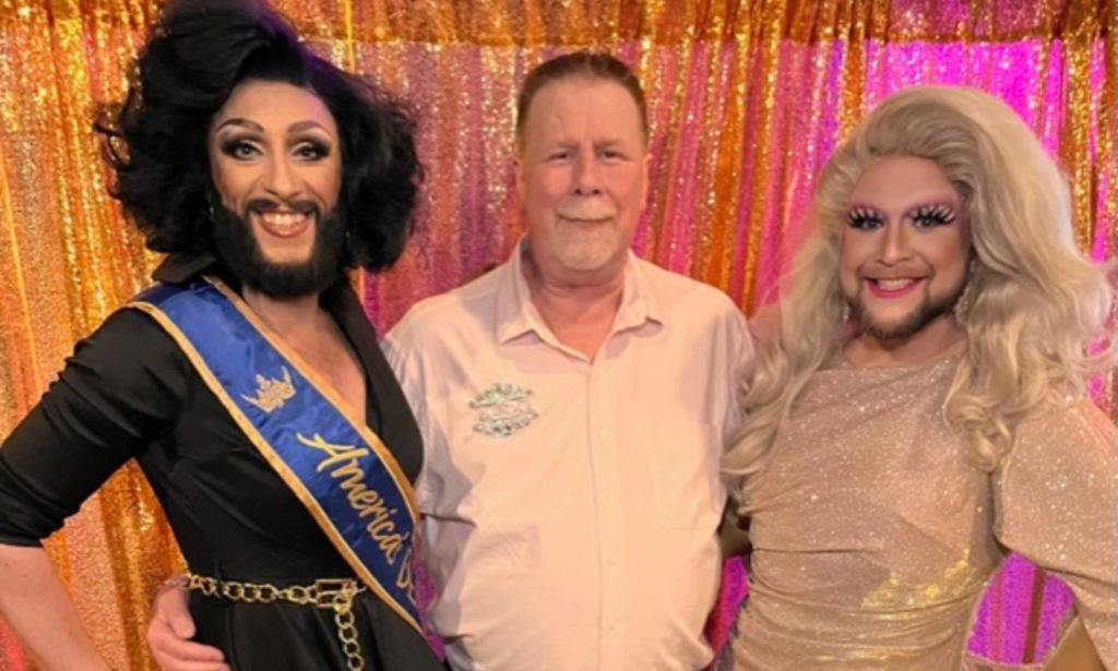 Rita Room wears a light blonde wig and sparkly dress as she stands alongside another bearded drag queen and the owner of Americas Bearded Queen Pageant System