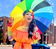 Tennessee drag queen Bella DuBalle, who has been fighting anti-LGBTQ+ bills and the state's drag ban, smiles while holding a rainbow umbrella. She's wearing an orange dress with rainbow accessories