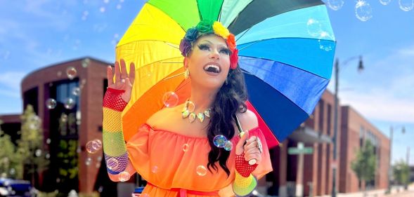 Tennessee drag queen Bella DuBalle, who has been fighting anti-LGBTQ+ bills and the state's drag ban, smiles while holding a rainbow umbrella. She's wearing an orange dress with rainbow accessories