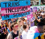 A crowd of people marching with a large trans flag with the words trans inclusion in schools