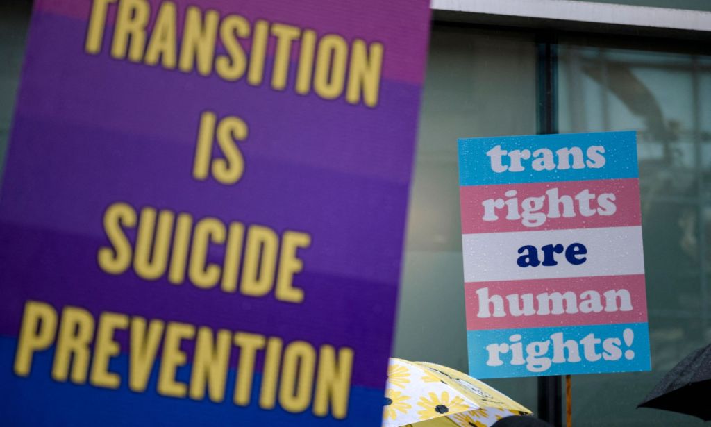 A person holds up a sign reading 'Transition is suicide prevention' while another holds up a sign reading 'trans rights are human rights' during a protest