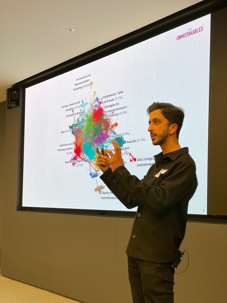 In this image, a man wearing black is speaking, behind him is a slide displaying graphical information.
