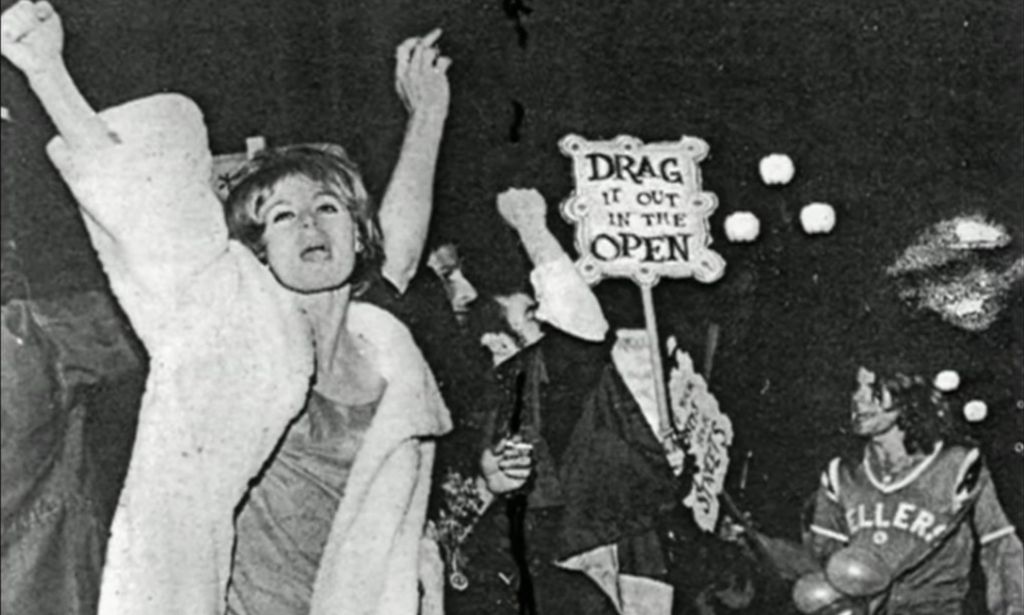 A photo from the Compton's Cafeteria Riots of various people protest police brutality against LGBTQ+ people and one person holds up a sign reading 'Drag it out in the open'