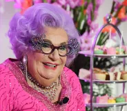 The late Barry Humphries dressed as his drag alter-ego Dame Edna Everage, with purple hair, horn-rimmed glasses and a pink dress