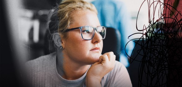 A female presenting person is looking out the window. They have eyeglasses on. They look stressed and there is a creative overlay for visual impact.