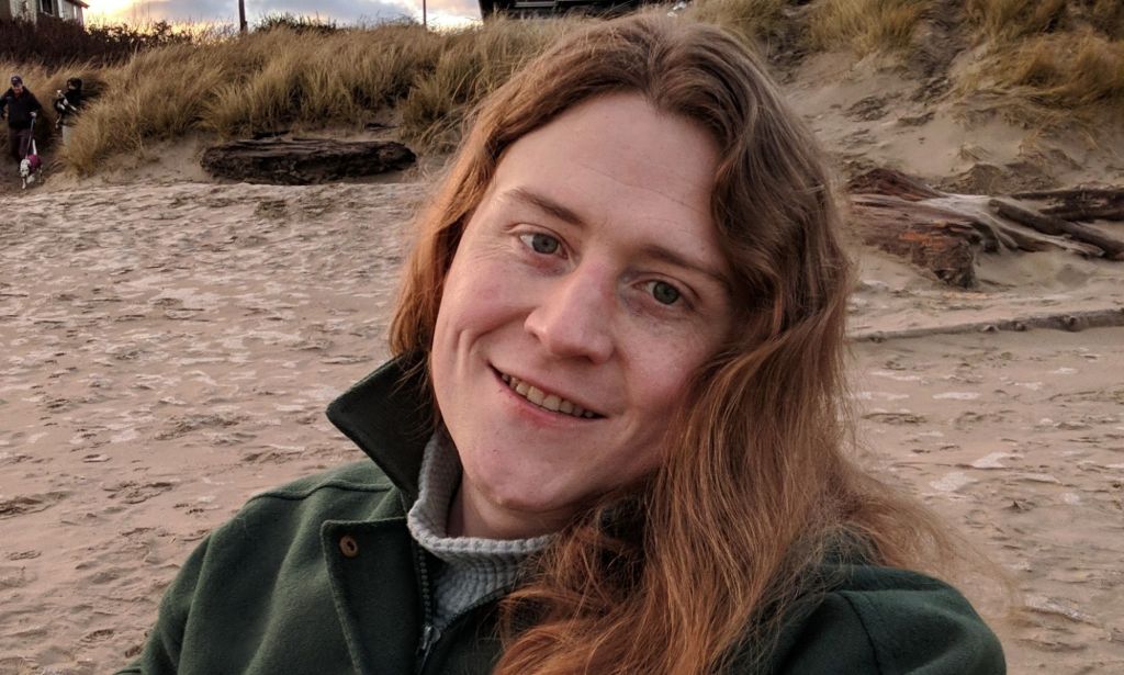 Non-binary American Ryan Castellucci wears a jacket and shirt as they chill on a sandy beach like setting