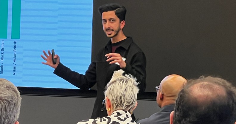 A man in a black shirt is speaking at an event whilst people in the audience look on.