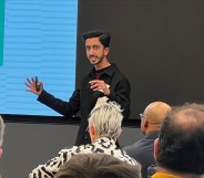 A man in a black shirt is speaking at an event whilst people in the audience look on.