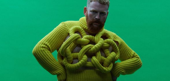Non-binary, trans singer Tom Rasmussen wears a yellow sweater while standing in front of a green background