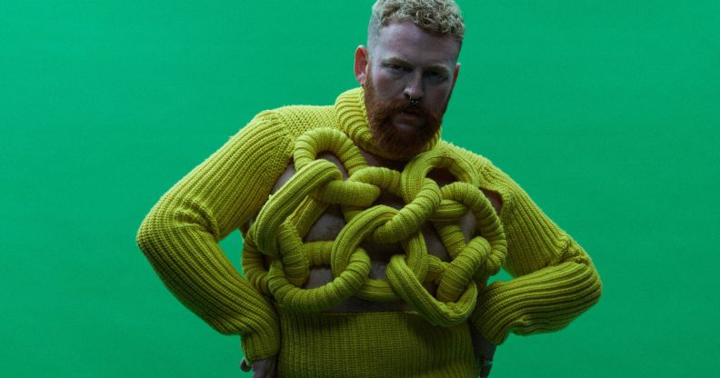 Non-binary, trans singer Tom Rasmussen wears a yellow sweater while standing in front of a green background
