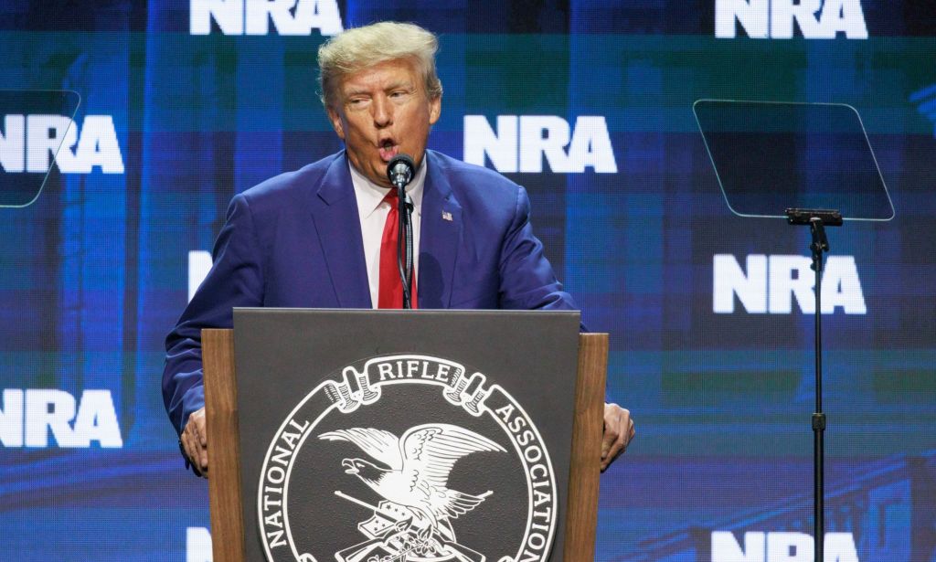 Former president Donald Trump wears a suit and tie as he stands at a podium and speaks into a microphone at the NRA annual conference