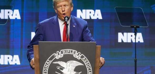 Former president Donald Trump wears a suit and tie as he stands at a podium and speaks into a microphone at the NRA annual conference