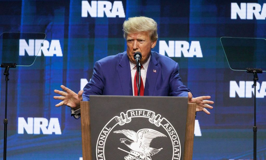 Former president Donald Trump wears a suit and tie as he stands at a podium and speaks into a microphone at the NRA annual conference
