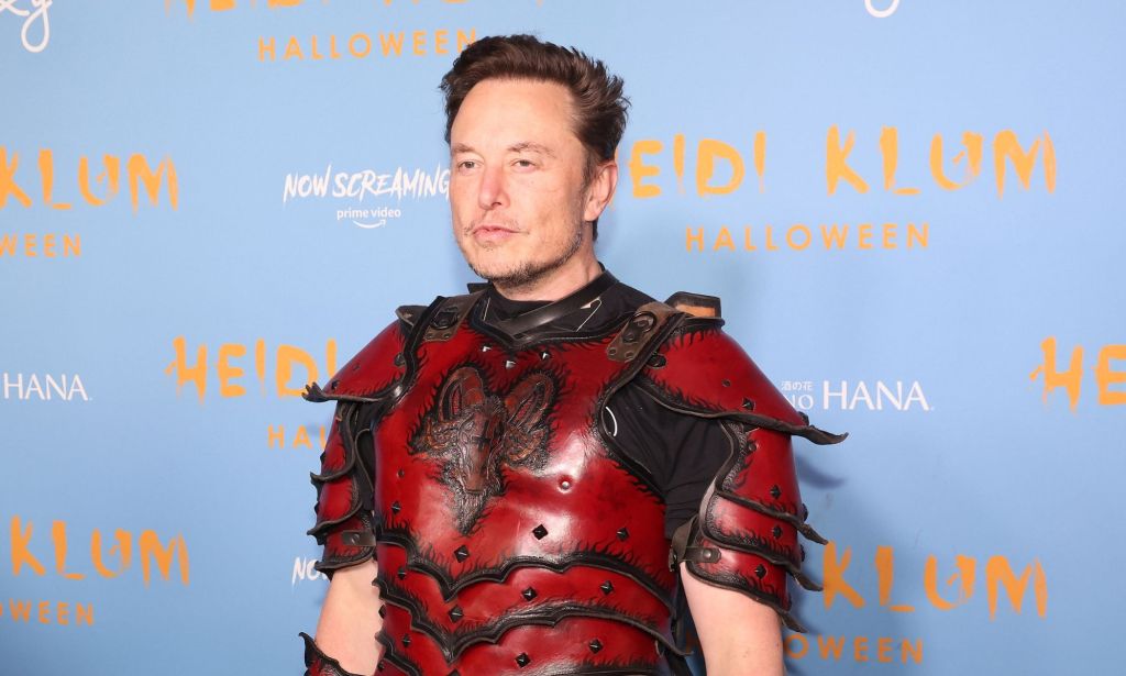 Twitter boss Elon Musk wears a red and black armour like outfit as he stands in front of a light blue and yellow background