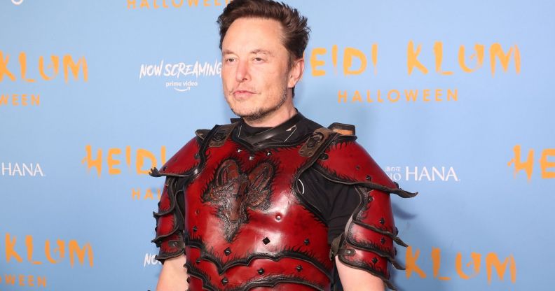 Twitter boss Elon Musk wears a red and black armour like outfit as he stands in front of a light blue and yellow background